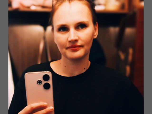 LisaLovHoney with fair skin and red hair, wearing a black shirt, holding a pink iPhone, taking a selfie with a slight smile, showing a small mole on her neck.