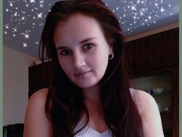 Close-up of LisaLovHoney with long, dark brown hair and blue eyes. She's wearing a white sleeveless top and small earrings. Her skin is fair with a slight pinkish tone. Sparkles are digitally added to the top of the image. She's looking directly at the camera with a slight, relaxed smile. The background shows a wooden cabinet with glass shelves. Her hair is slightly messy, adding a casual, everyday feel.