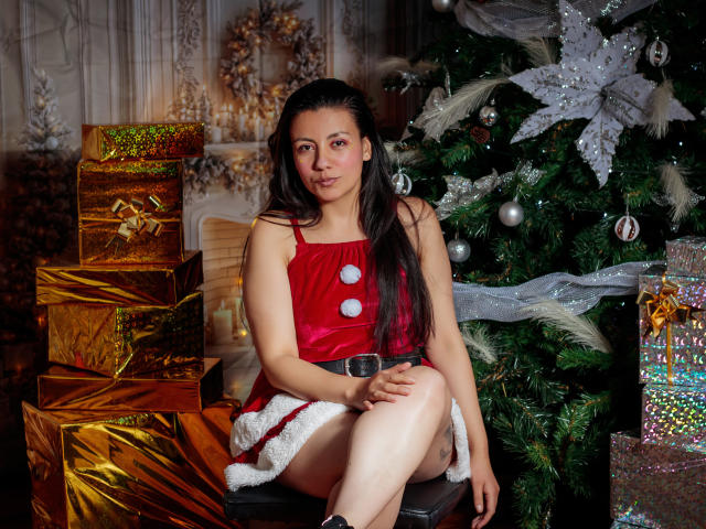 MargarettCooper with long dark hair, red velvet dress with white pom-poms, sitting by a Christmas tree, surrounded by gold and glittery wrapped gifts, looking cute and relaxed.