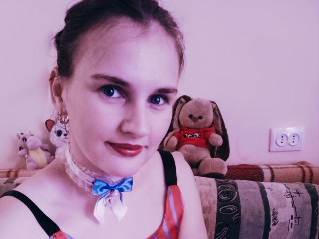 LisaLovHoney with blue eyes, red lipstick, white lace choker with blue ribbon, pink and black dress, sitting next to stuffed bunnies and cats on a striped couch.