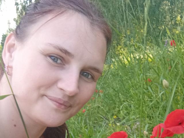 LisaLovHoney's face with blue eyes, light skin, brown hair, slight smile, wearing small earrings, in a grassy field with red and yellow flowers in the foreground.