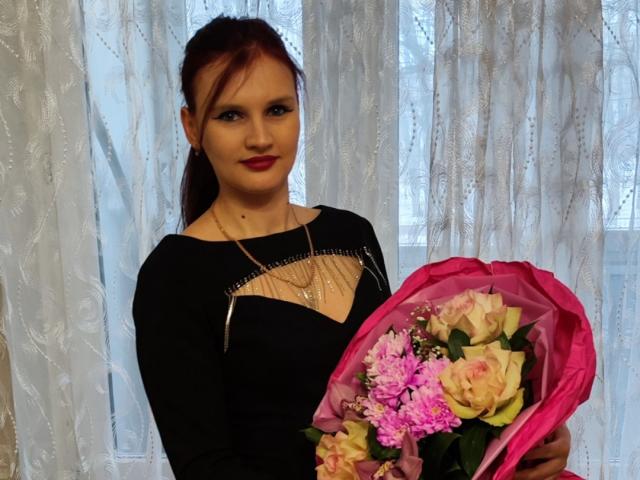 LisaLovHoney with red hair, black dress, red lipstick, holding pink bouquet of roses and pink carnations, standing in front of white lace curtain, holding bouquet in both hands.