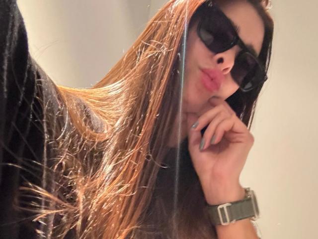 Pupita-hot with long brown hair, black sunglasses, and glossy pink lips, pouting while touching her chin. Wearing a black shirt and green watch.