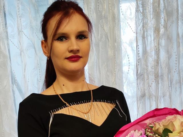 LisaLovHoney with red hair, black dress, sheer neckline, gold necklace, holding pink flowers, has red lipstick and green eyes, standing in front of white lace curtains.