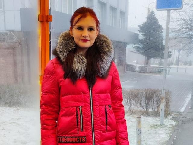 LisaLovHoney in a red puffy jacket with a furry collar, standing indoors, snow outside, red lipstick, red hair, slightly smiling, "THEOQUITS" logo on jacket.