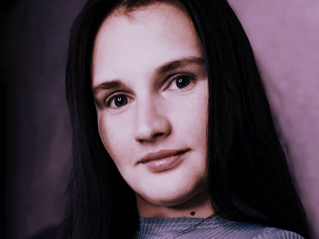 Close-up of LisaLovHoney with pale skin and dark, straight hair. She has brown eyes, a small black mole under her right cheek, and a subtle, calm expression. Her lips are slightly parted, and she's wearing a gray, textured top. The image has a purple tint, and the background is blurred, keeping focus on her face. Her eyebrows are dark and slightly arched. The texture of her hair is smooth, and the overall composition is centered.