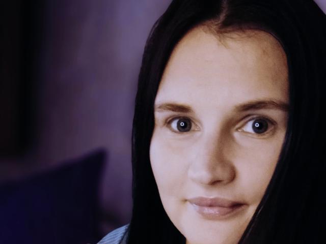Close-up of LisaLovHoney's face. She has fair skin, dark brown straight hair, and blue eyes. Her eyebrows are thin and slightly arched. She has a subtle, almost neutral smile. The lighting is soft, highlighting her facial features. The background is blurred and dark, with a hint of purple and blue. Her hair is parted in the middle. The image is sharply focused on her face, with a slight depth of field that blurs the background.