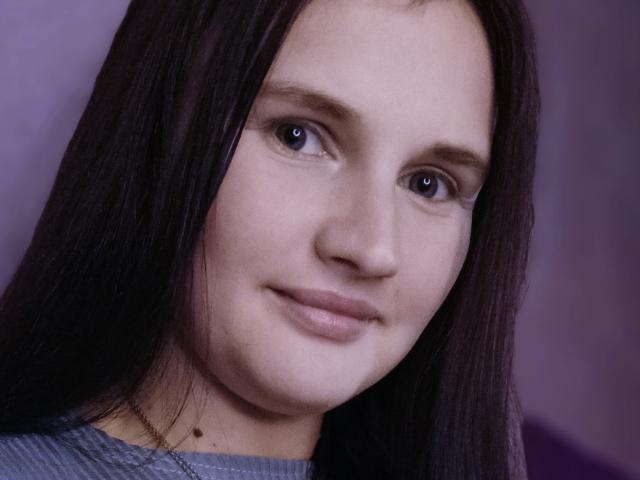 LisaLovHoney has pale skin, dark straight hair, small brown eyes, and a slight smile with no makeup. She's wearing a gray shirt, showing a small mole on her chin.