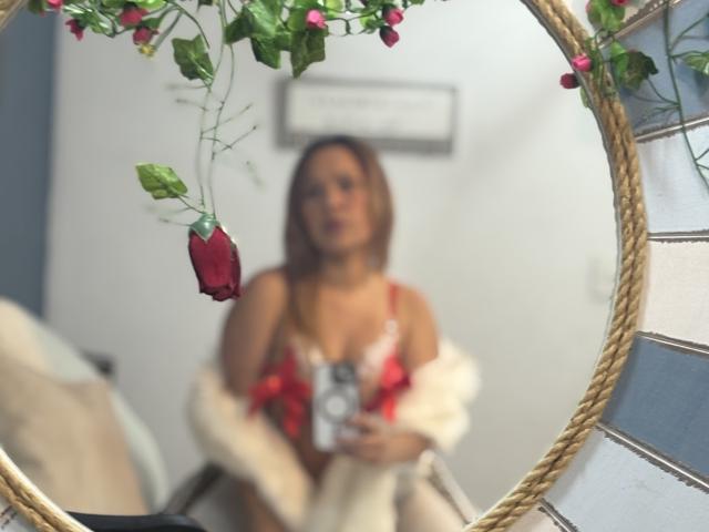 Reflection in a round mirror shows SophiaQueens with red rose petal in her hair, wearing red and white lingerie, holding a camera, and draped in a white fur.