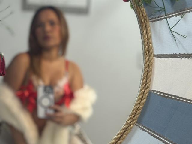Blurred mirror reflection of SophiaQueens with red floral bra, white off-shoulder sweater, holding phone, rope decor, and striped wall. Boobs slightly visible, face out of focus.