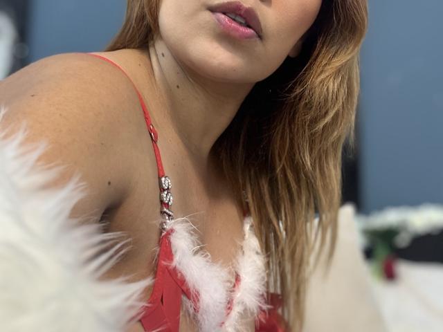 SophiaQueens with light brown hair, pink lips, and red lace bra with white fur trim, showing her small tits and slightly tanned skin. Blurred background.