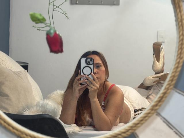 SophiaQueens lying on a bed, taking a selfie with her phone, wearing a red top, white shorts, and beige heels, with a rose in the mirror's reflection.