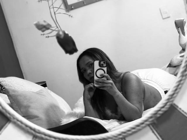 SophiaQueens with long hair, lying on bed, taking mirror selfie with phone, wearing white underwear, showing small tits, looking at camera, in black-and-white photo.