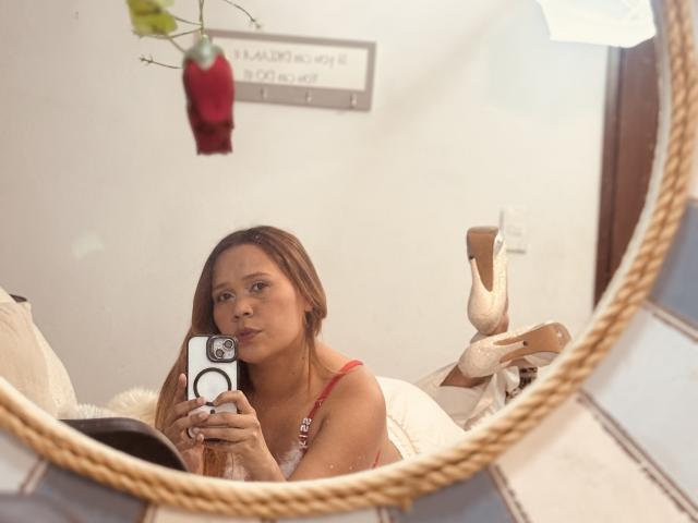 SophiaQueens with brown hair, red bra, and a cute smile, taking a mirror selfie with her phone, lying on a white couch, wearing beige high heels, and a red rose.
