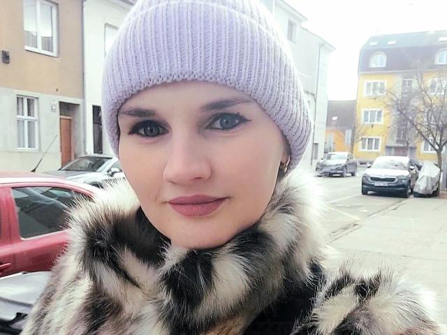 LisaLovHoney with pale skin, blue eyes, and pink lips, wearing a lavender knit beanie and a fur scarf, standing in a snowy urban area with parked cars.