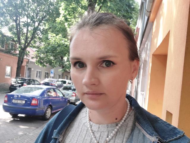 LisaLovHoney with a serious face, blue eyes, and a small mole on her chin, wearing a pearl necklace and denim jacket, standing in a street with parked cars.