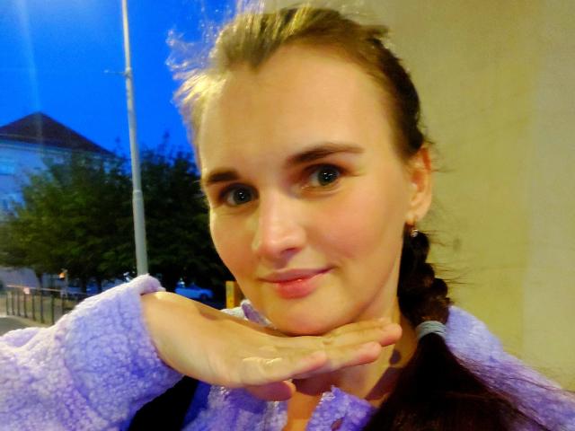 LisaLovHoney with her hand under her chin, smiling slightly, wearing a purple sweater, blue eyes, brown hair in a braid, fair skin, small earrings, against a dimly lit background.