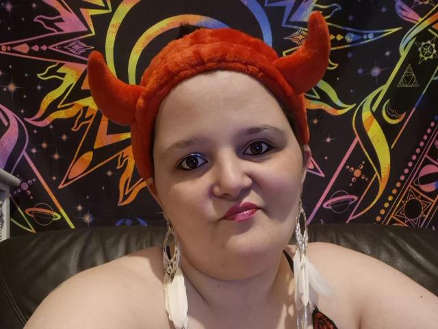 Kimy69 with red devil horns, chubby face, pink lipstick, black eyeliner, white feather earrings, and a brown leather chair. She's smiling slightly, wearing a red velvet headband.