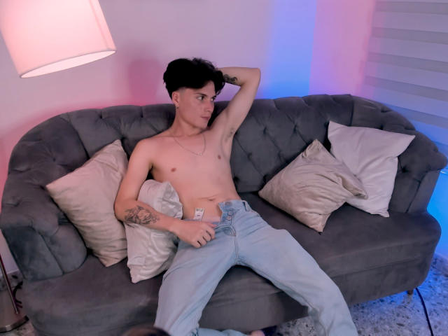 Jack Rouse sin camisa, con un tatuaje en el brazo izquierdo, vistiendo jeans azul claro, recostado en un sofá gris con almohadas blancas, sosteniendo un condón en su mano derecha..