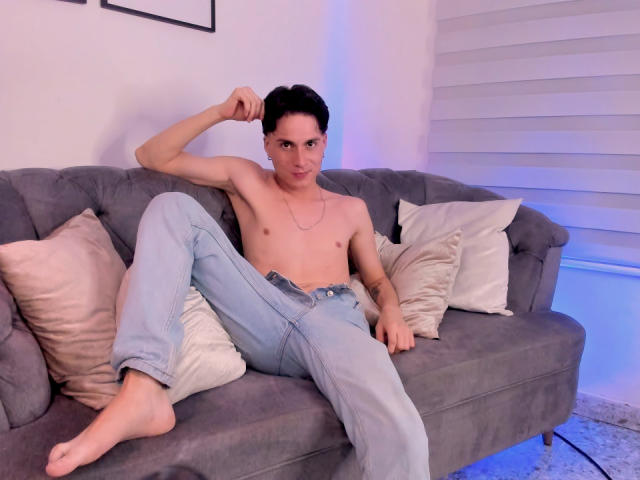 JackRouse, sin camisa, con un cuerpo delgado y tonificado, vistiendo jeans azul claro, sentado en un sofá gris, con las piernas separadas, el brazo derecho levantado, mirando a la cámara, con tatuajes en el brazo izquierdo..