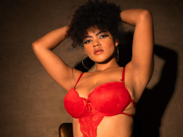 AliceJelanni with curly black hair, wearing red lace bra, arms raised, looking at camera, large breasts, hoop earrings, brown skin, gold makeup, sexy, warm lighting, shadow on wall.