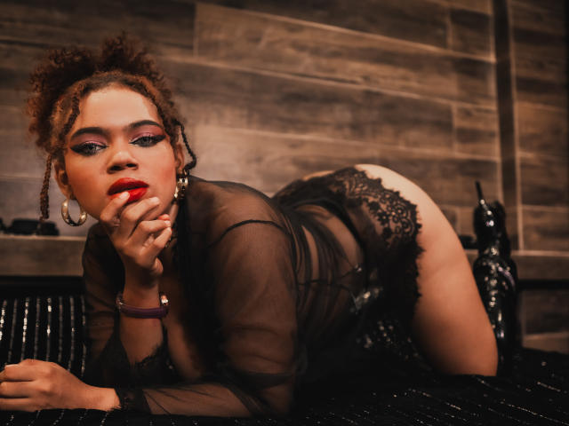 AliceJelanni on all fours, wearing sheer black lingerie, lace panties, and red lipstick, with curly brown hair, black boots, and purple bracelet, sucking her finger, looking at camera.