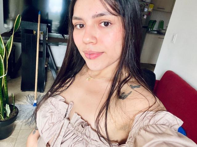 SophiaMarx with long dark hair, light brown skin, and pink lips, wearing a pink off-shoulder top, showing small tits and tattoos on her right shoulder. She's smiling softly.