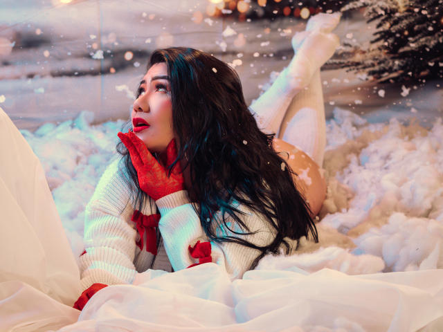 KhloeLawson laying in snow, wearing white sweater, red gloves, white thigh-high socks, red lipstick, long black hair, looking up, butt raised, hands near face, snowy background.
