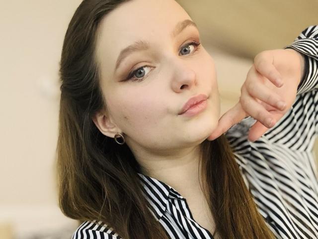 MagikPeachh has light skin, brown hair, and blue eyes. She's wearing a black and white striped shirt, with her right hand touching her chin. She has subtle pink lip color, light brown eyeliner, and small hoop earrings. Her hair is straight and parted to the side. The composition focuses on her upper body and face, with a slight upward angle. The image has a soft, natural light. Her expression is neutral, slightly curious. The overall style is casual and modern.