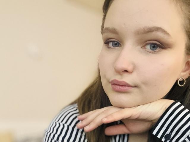 MagikPeachh's face, light skin, blue eyes, small nose, pink lips, brown hair, wearing striped shirt, hand under chin, gold hoop earring, slightly smug expression.
