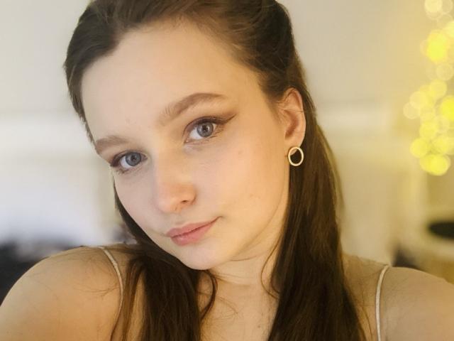 MagikPeachh has pale skin, blue eyes, and brown hair, wearing small hoop earrings, with a subtle smile, and is showing her tits in a soft-focused selfie.