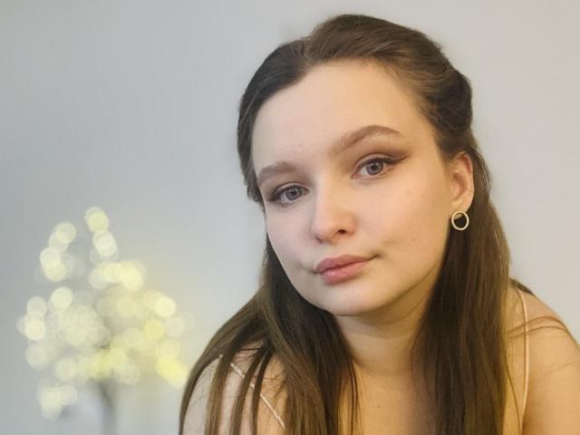 Close-up of MagikPeachh with fair skin and long brown hair. She has small hoop earrings, subtle eyeliner, and pinkish lips. Her expression is neutral, slightly pouty. She wears a light-colored top. The blurred background has bokeh light effects. The image has a soft, natural light. MagikPeachh's face is centered, and her hair is parted in the middle. The focus is on her facial features and expression.