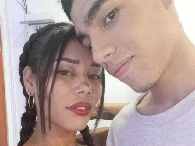 Ethanica69 with red lipstick, braided hair, hoop earrings, nose ring, leaning on a guy with short dark hair, light skin, gray shirt, and a slight smile.