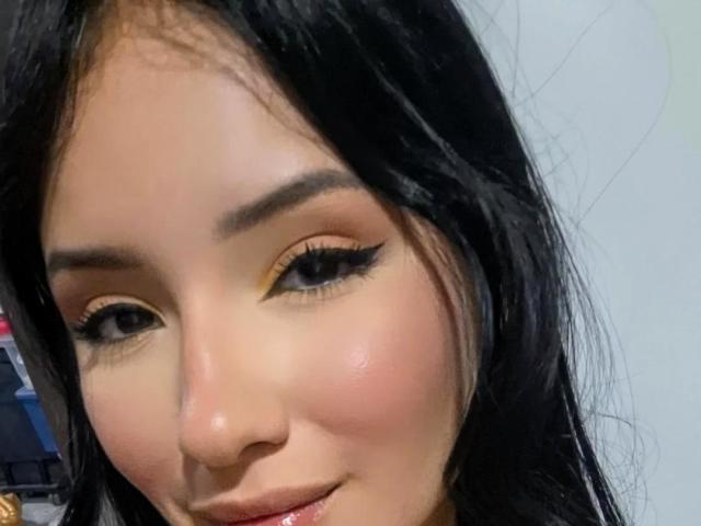 LizzJoy has tan skin, black eyelashes, orange-yellow eyeshadow, and a subtle smile. Her long black hair is slightly messy. She's showing off her cute, round ass-face.