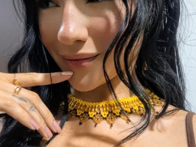 LizzJoy's face with black wavy hair, wearing a yellow beaded necklace, touching her chin with a hand showing a tattoo and gold ring, slightly smiling.