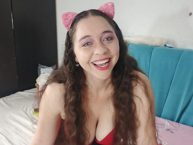 NalimiSmirnov, smiling with pink cat ears, has long curly brown hair, purple eyeshadow, and is wearing a red dress that shows her big boobs. She's on a bed.