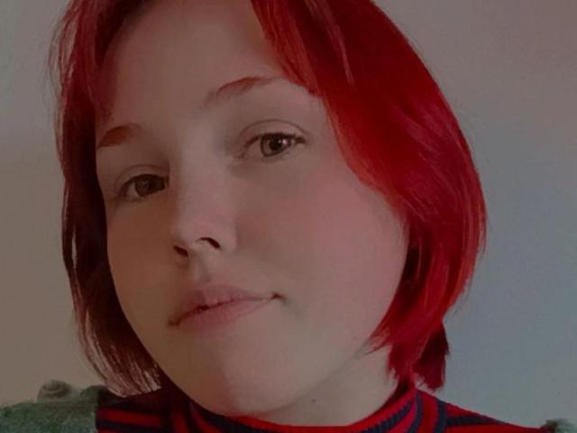 EvelinaRedFoxy has red hair, light skin, and is looking at the camera with a slight smile, wearing a red and black striped shirt, showing her cute face.