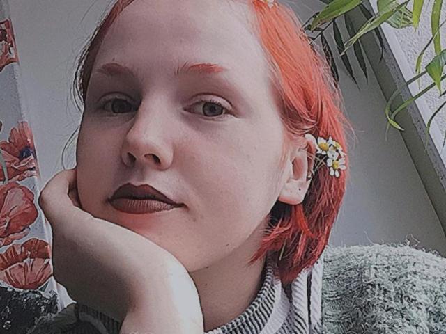 EvelinaRedFoxy has pale skin, red hair, and wears a flower in her ear. Her hand rests on her chin, with dark red lipstick and a subtle smile.