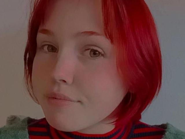 EvelinaRedFoxy with red, shoulder-length hair, light skin, brown eyes, slight smile, wearing striped shirt, green sweater, subtle makeup, soft lighting, slight blush on cheeks, relaxed expression.
