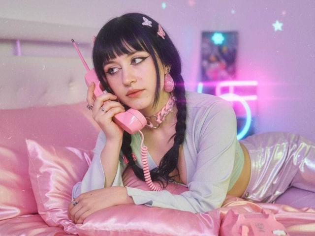 Velmoon avec un téléphone rose, portant des tresses, un collier rose, un haut gris clair, un pantalon argenté brillant, allongée sur un lit rose, tenant un téléphone à l'oreille, l'air ennuyée, mignonne, pastel, kawaii..