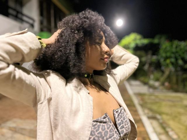 Megan Williams, avec ses cheveux noirs bouclés, portant une veste beige et un haut à imprimé léopard, les mains dans les cheveux, nuit éclairée par la lune, bracelet vert, veste légèrement ouverte laissant entrevoir sa poitrine..