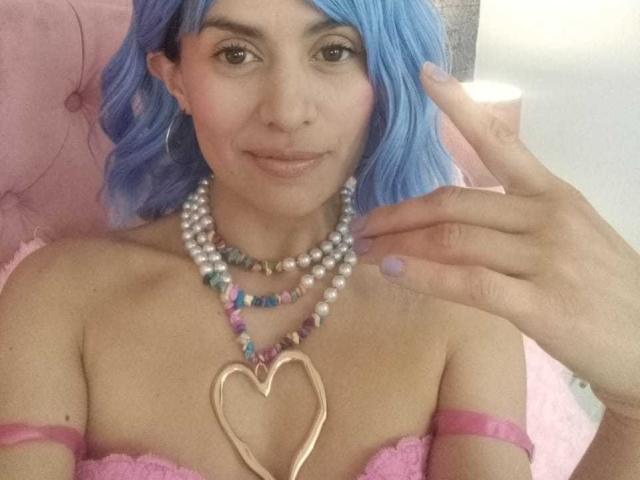 LilaaSalmon with blue hair, wearing pink lace top, pearl necklace, multicolored bead necklace, heart-shaped pendant, pink arm bands, touching her chin, smiling, showing her tits.