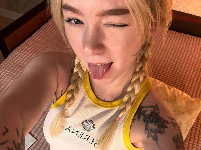 EnchantingEyes with blonde braids, winking and sticking out her tongue, wearing a yellow-trimmed tank top, showing tattoos on her arm and upper chest, in a selfie.