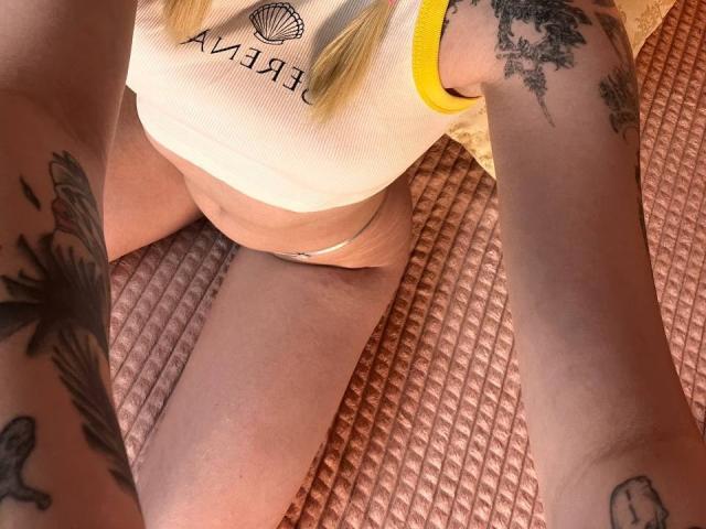 EnchantingEyes with tattoos on arms, wearing white ribbed shirt with yellow trim, and white panties, sitting on pink textured mat, showing thighs and tattoos, taken from above.