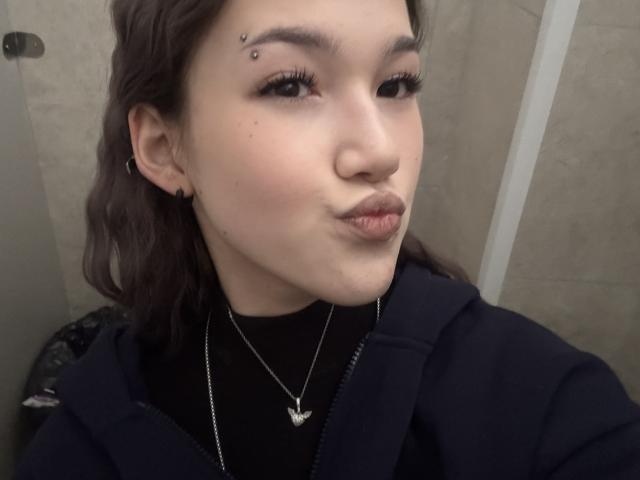 LixDomii with dark brown hair, black eyeliner, and a small black beauty mark above her right eyebrow, pouting her lips. She's wearing a black top and a silver necklace.