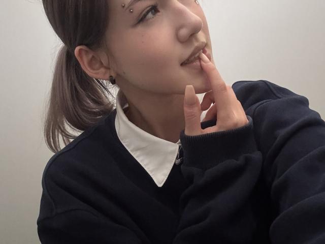 LixDomii has brown hair in a ponytail, a black sweater, white shirt, and black ear studs. She's biting her finger with small facial piercings on her upper lip.