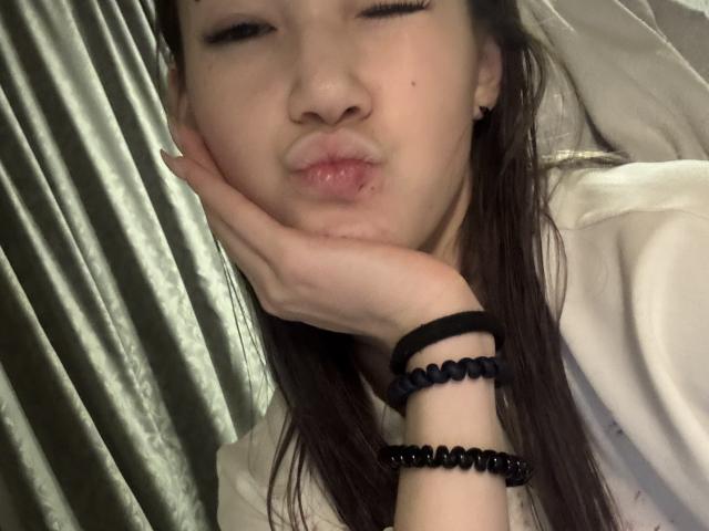 LixDomii's face is puffed up like a fat ass, with her hand on her cheek, wearing black bead bracelets, and her eyes are winking. She's got long brown hair.