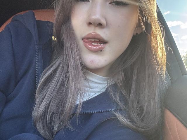 LixDomii's face with shiny pink lips, slightly parted, wearing a blue hoodie, white shirt, and brown car seat. She has light brown hair, and a small pimple on her chin.