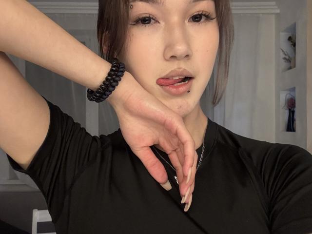 LixDomii with a black shirt, tongue slightly out, showing a lip piercing, left hand near chin, black bracelet, and long nails, looking at camera with a subtle expression.