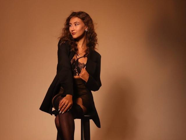 A photo of Afinaa with curly brown hair, sitting on a black stool against a beige background. She wears a black blazer, sheer black top, and black fishnet stockings. Afinaa's left hand rests on her thigh, right hand holds the blazer's lapel. She looks to the side with a slight, confident smile. Soft shadows add depth. Afinaa's skin is a warm brown, and her earrings are small and metallic. The overall composition is simple and elegant.